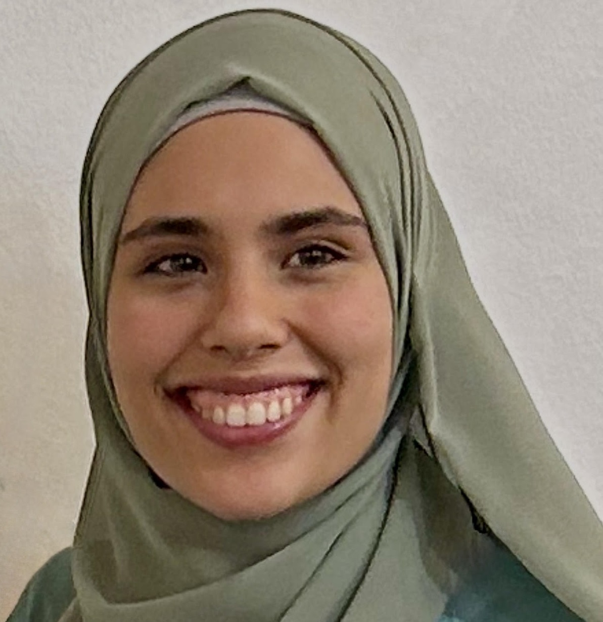 a headshot photograph of Aisha Benloucif. She is wearing a grey scarf and blue outfit, posing with a smile on her face in front of a white background