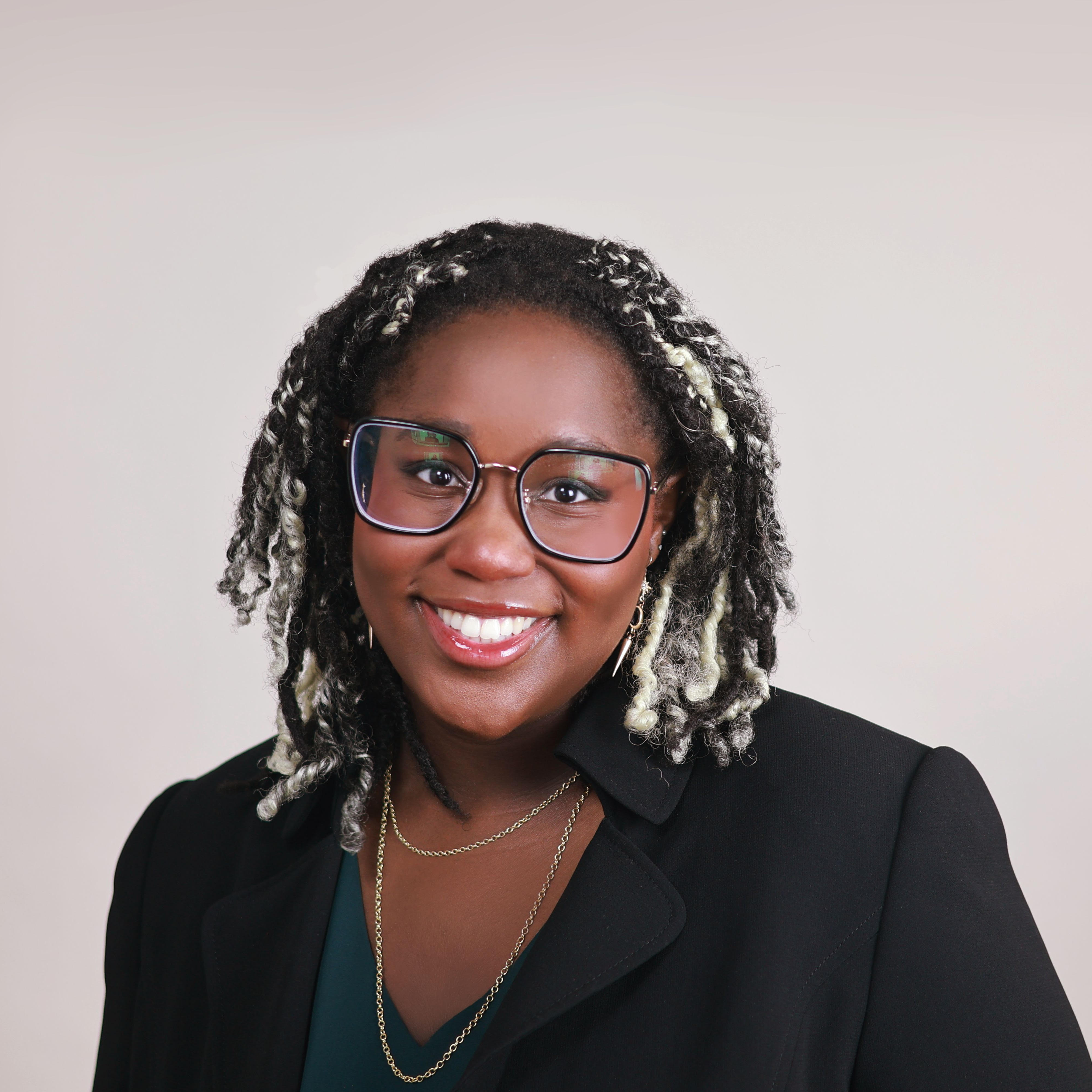 a studio headshot of leah smiling, with a platinum blond and black twist hairstyle, wearing a blazer and green blouse