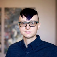 A headshot of Luke, a white person with short, dyed purple hair wearing a dark blue buttoned shirt and turquoise-rimmed glasses. In the background, there is blurred abstract art against an off-white wall.