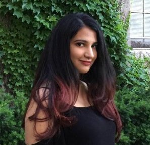Rahaf with dyed maroon hair tips. In the background, there are green plants hanging on the wall. Rahaf is wearing a black t-shirt
